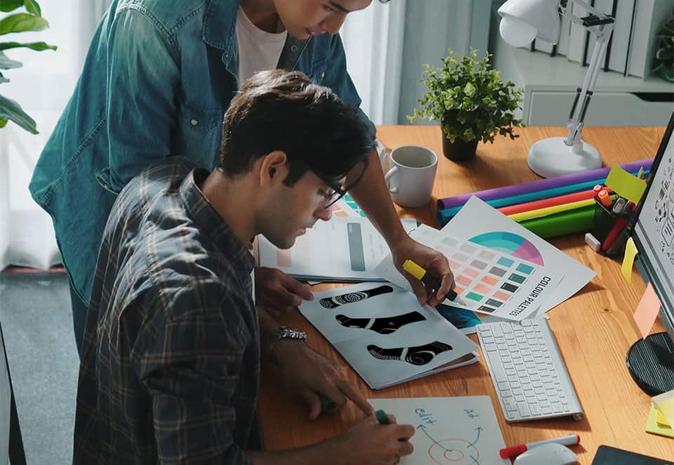 Two people work at a desk with color swatches, a computer, shoe design printouts, pens, a lamp, and a potted plant.