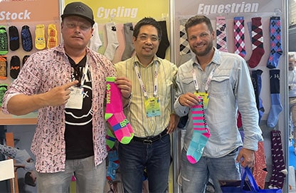 Three men at a "Cycling" and "Equestrian" trade booth hold colorful socks in front of a sock display.