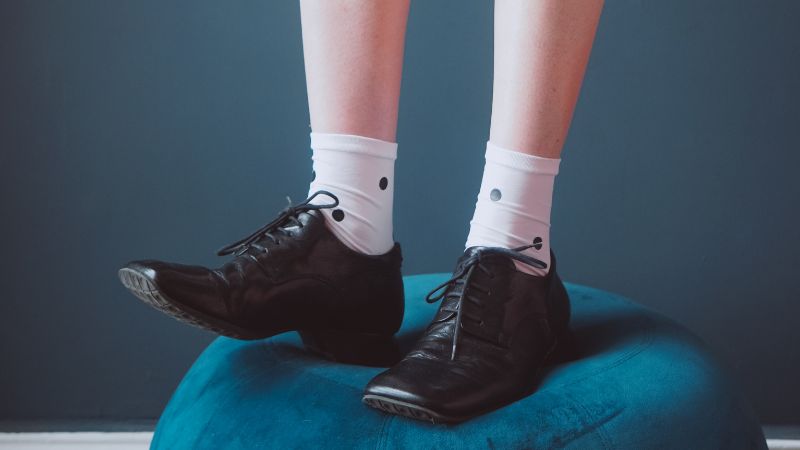 Legs in Sinoknit white socks with black dots and loafers rest on a blue cushion against a dark teal background.