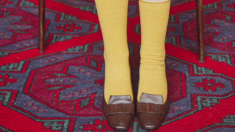 Someone in yellow Sinoknit socks and brown loafers stands elegantly on a colorful rug.