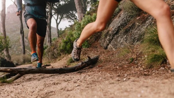 Dos personas corriendo por un sendero forestal rocoso.
