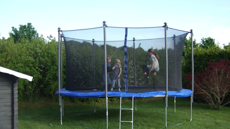 Tiga anak dengan gembira melompat di atas trampolin yang aman dengan jaring pengaman, terletak di halaman belakang yang dikelilingi pepohonan.