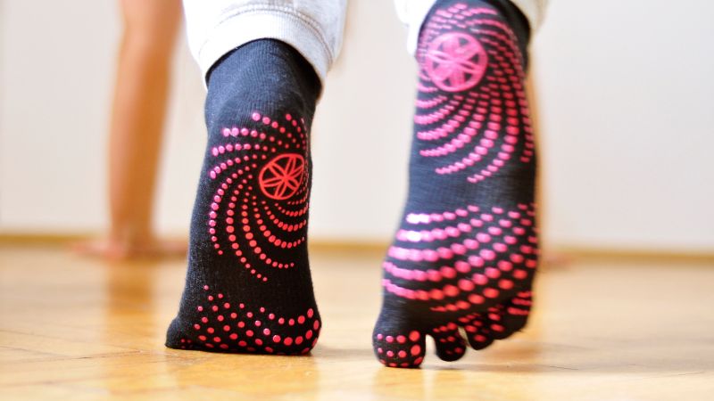 Une personne portant des chaussettes noires à pois roses se tient debout sur un plancher en bois. Pour laver facilement des chaussettes antidérapantes et les garder éclatantes, suivez ces étapes.