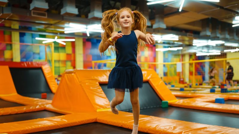 A girl with long hair joyfully jumps on a trampoline indoors, highlighting the importance of trampoline safety for kids.