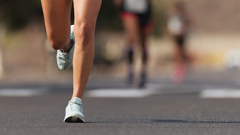 Close-up of a jogger on a sunlit road in Merino Wool Socks, with stylish athletic shoes and another runner blurred in the background.