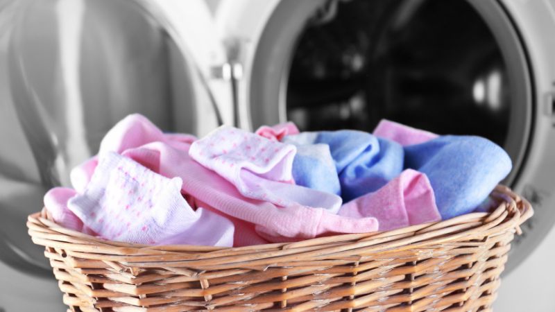 Un panier en osier contenant des vêtements roses et bleus, y compris des chaussettes antidérapantes, se trouve devant une machine à laver ouverte.