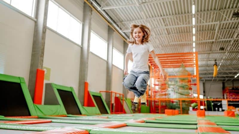 A child in a white shirt and gray pants jumps on a trampoline inside an indoor park, wearing trampoline socks.