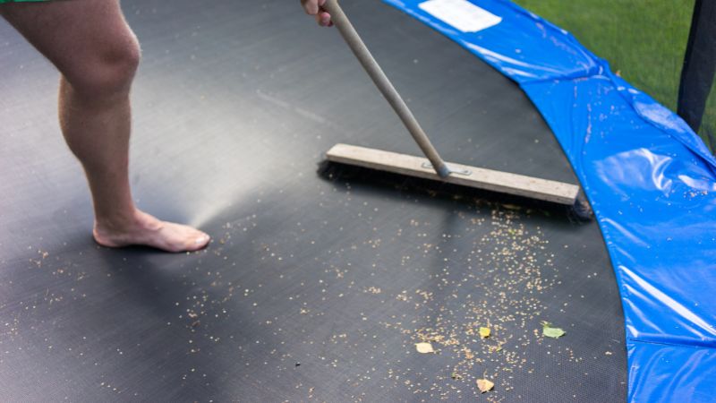 Close-up of person sweeping debris off a backyard trampoline with a broom, highlighting maintenance and cleaning services for outdoor trampoline safety and longevity.