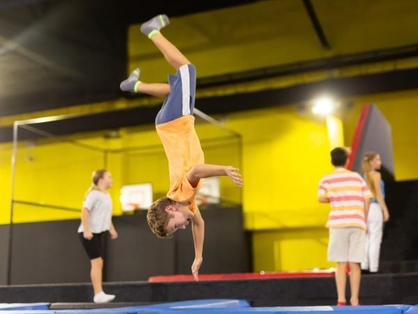 Young girl performing back handspring at indoor trampoline park or gymnastics facility with teammates in background.